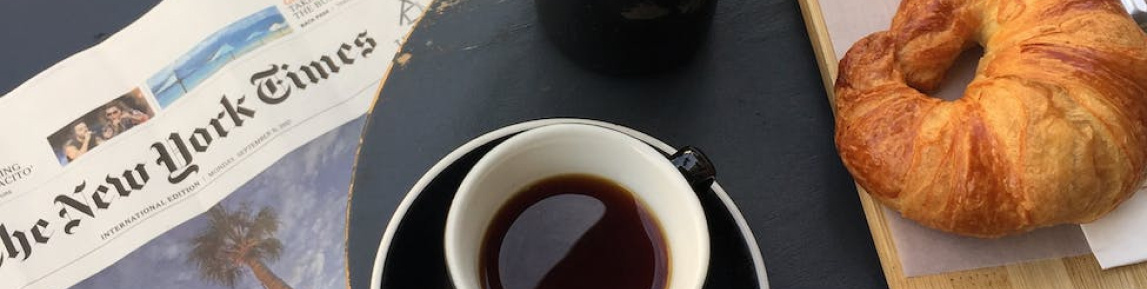 A coffee, croissant, and newspaper on a table.