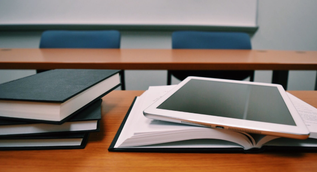 Two stacks of books atop a wooden desk in classroom setting